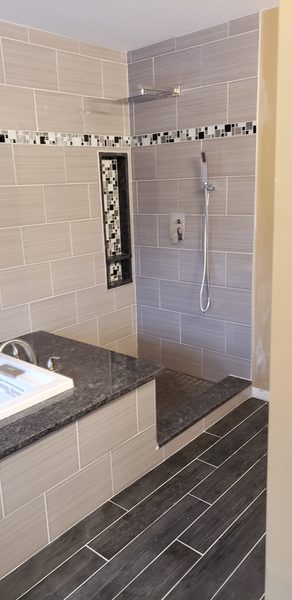 Walk-in shower in linear gray tile with a glass mosaic accent band and niche, beside a granite tub deck with a drop-in tub