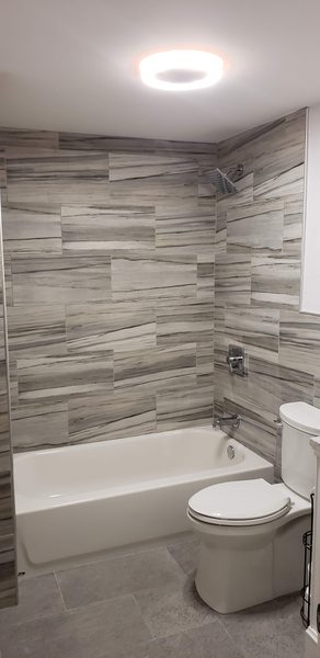 Tub and shower alcove in gray marble-look plank tile with a rain shower head, white tub and toilet